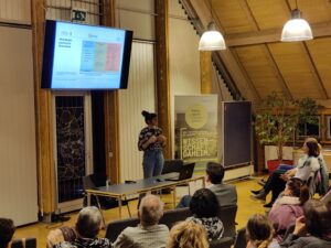Eine Frau, Christine Preiser, hält einen Vortrag vor einem Heimspiel. Ein Roll-Up steht im Hintergrund, auf dem steht "Heimspiel Wissenschaft". Vor ihr sitzt ein gesmichtes Publikum und hört gespannt zu. 